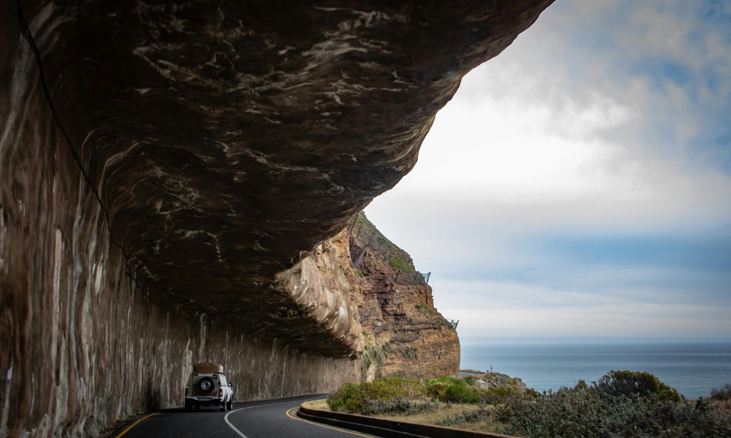 SUV driving along a coastal road under a rocky overhang in Cape Town, South Africa.